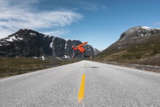 A traveler in a red jacket leaps mid-air on an open mountain road in Norway Framed by dramatic