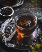 A warm glass cup of tea infused with anise, set on an intricately designed metal tray. The aromatic