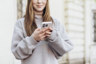 Teenage girl holding a smartphone while outdoors, dressed in a casual gray sweatshirt. The focus is