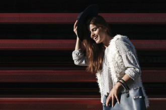 A fashionable woman poses outdoors on a sunny day, wearing a black hat, patterned shirt, and fur