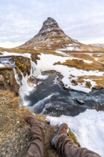 POV of a man sitting on the waterfall with snow on the famous Kirkjufell mountain in winter Iceland