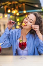 Vertical photo of a caucasian woman enjoying biting the citrus garnishing a cocktail in an outdoor