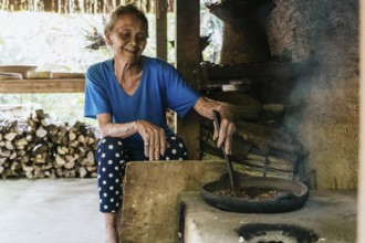 An elderly woman in a blue shirt sits by a rustic stove, stirring a frying pan with balinese coffee