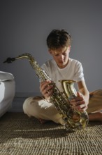A young musician sits indoors in soft natural light, examining a saxophone. The scene captures