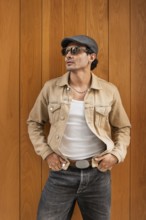 A fashionable man wearing sunglasses, a tan jacket, and a cap stands confidently against a wooden