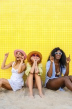 Three diverse women wearing summer clothes and hats are sitting on the sand, pointing upwards with