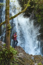 A man stands on a rock overlooking a waterfall. He is wearing a backpack and a blue hat. The scene