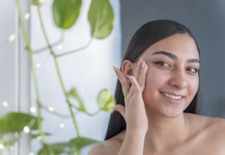 Young woman with healthy, glowing skin smiling gently, caressing her face in a serene setting with