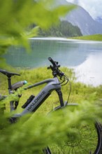 An e-bike is parked on the lakeshore. The surrounding nature is lush and green, and the mountains