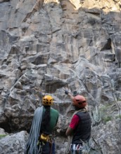 Two climbers equipped with helmets and ropes assess a towering rock face for a traditional climbing
