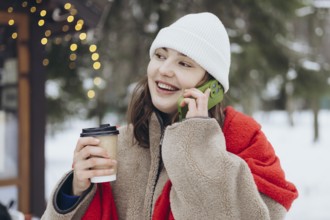 A young woman dressed in winter clothing holds a coffee cup and smiles while chatting on her phone
