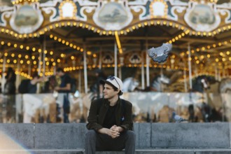 A young man sits casually in front of a brightly lit carousel in Brooklyn Bridge Park. Pigeons fly