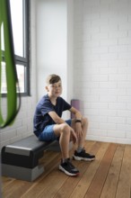 A young boy sits on a bench in a modern, brightly lit room with wooden floors and white brick