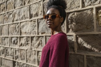 A stylish woman in a burgundy sweater wearing sunglasses stands confidently against a textured