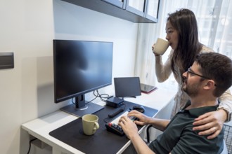 A diverse couple is sharing a cozy moment at home. One is working on a computer while the other