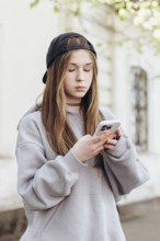 Teenage girl in casual clothing, engrossed in her smartphone while standing outdoors. She appears