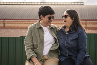 A joyful curvy lesbian couple shares a light heart moment outdoors, sitting closely and wearing