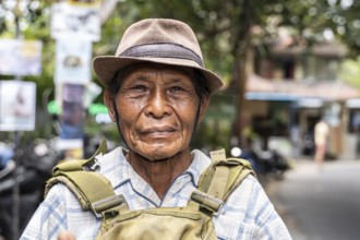 An elderly balinese man wearing a hat and a plaid shirt stands outdoors, looking into the camera
