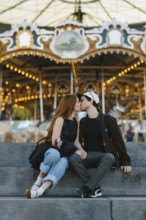 A couple shares a kiss on stone steps in front of a brightly lit carousel in Brooklyn Bridge Park.