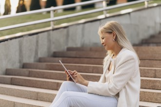 A businesswoman in casual business attire sits on outdoor steps while using a tablet. The setting