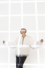 A middle-aged woman with a shaved head poses confidently in a chic white fur coat and stylish pants