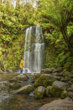 A woman stands by a waterfall in a lush forest, surrounded by moss covered rocks and vibrant ferns,