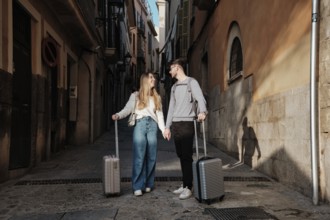 A young couple walks hand in hand down a narrow street, luggage in tow, as they search for their