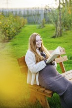 Beautiful pregnant woman with long red hair enjoys a serene moment on a park bench, reading a book.