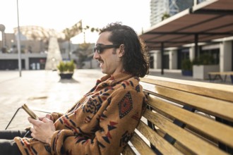 A man sits calmly on a bench outdoors with a smartphone Wearing sunglasses and a stylish patterned