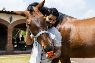 A man affectionately feeds his horse from a red container while embracing it on a sunny day at the