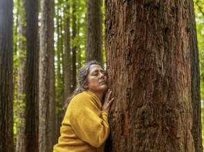 A woman in a yellow sweater embraces a tall tree, eyes closed, connecting deeply with nature in a