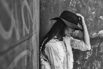 A black and white image of a woman in a hat resting against a textured wall. She wears a stylish