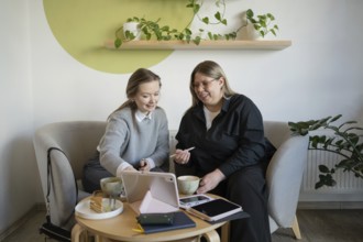 Two women sit on cozy chairs in a cafe, sharing conversation over coffee. A tablet rests on the