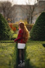 A young woman with long red hair explores a lush green maze in a Montreal garden during fall.