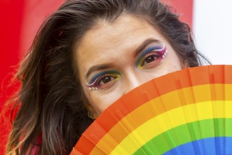 A woman with vibrant rainbow makeup partially conceals her face with a pride-themed fan. Her