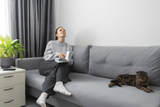 A woman sits on a gray sofa, holding a book, looking peaceful and contemplative. Beside her, a cat