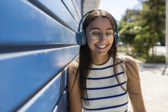 Young woman enjoying her urban lifestyle outdoors, leaning against a blue wall while listening to
