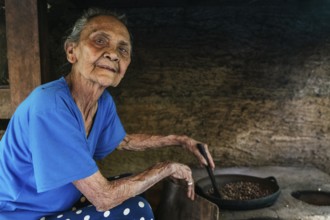 An elderly woman in a blue shirt sits by a rustic stove, roasting balinese coffee beans in a pan.