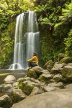 A woman sits on rocks, meditating beside a serene waterfall, surrounded by lush greenery in the