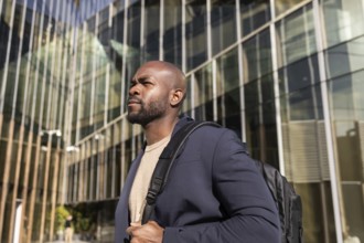 A confident cuban man with a backpack stands outside a modern office building. The glass facade