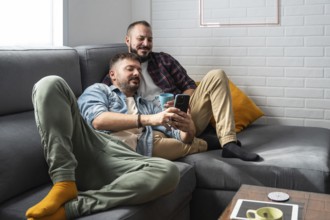 A content gay couple lounges on a comfortable gray sofa, enjoying coffee together while browsing a