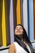 A woman in a colorful outfit enjoys the sunlight, standing in front of a backdrop of vibrant,