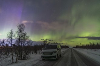 A mesmerizing display of Aurora Borealis fills the sky above a snowy road with a vehicle parked