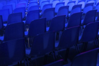 A series of empty blue chairs are arranged in neat rows under a dramatic blue spotlight at a