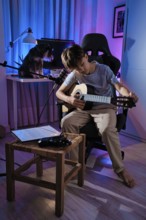 A young boy tuning his guitar in a cozy, blue lit room, with a microphone and sheet music nearby. A