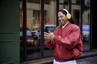A man in a red jacket, wearing yellow headphones, smiles while looking at a smartphone. Captured