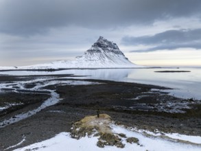 An unidentified man stands on a snow-dusted outcrop facing Kirkjufell, Iceland's iconic mountain,