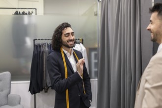 A tailor communicates with a customer in a suit store, showcasing a friendly ambiance. Suits hang