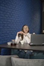 A woman with glasses sits at a cafe table with a laptop, tea cup, and teapot, against a stylish