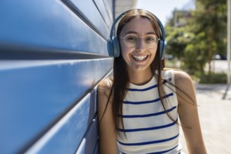 Smiling young woman with headphones enjoys music while leaning against a blue wall in an urban